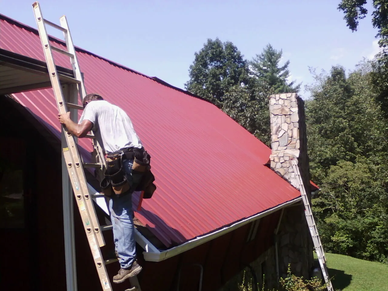 Roofer on a ladder beside a red metal roof and stone chimney — Soffit & Fascia Repair by Roofing Brothers in Fort Monroe